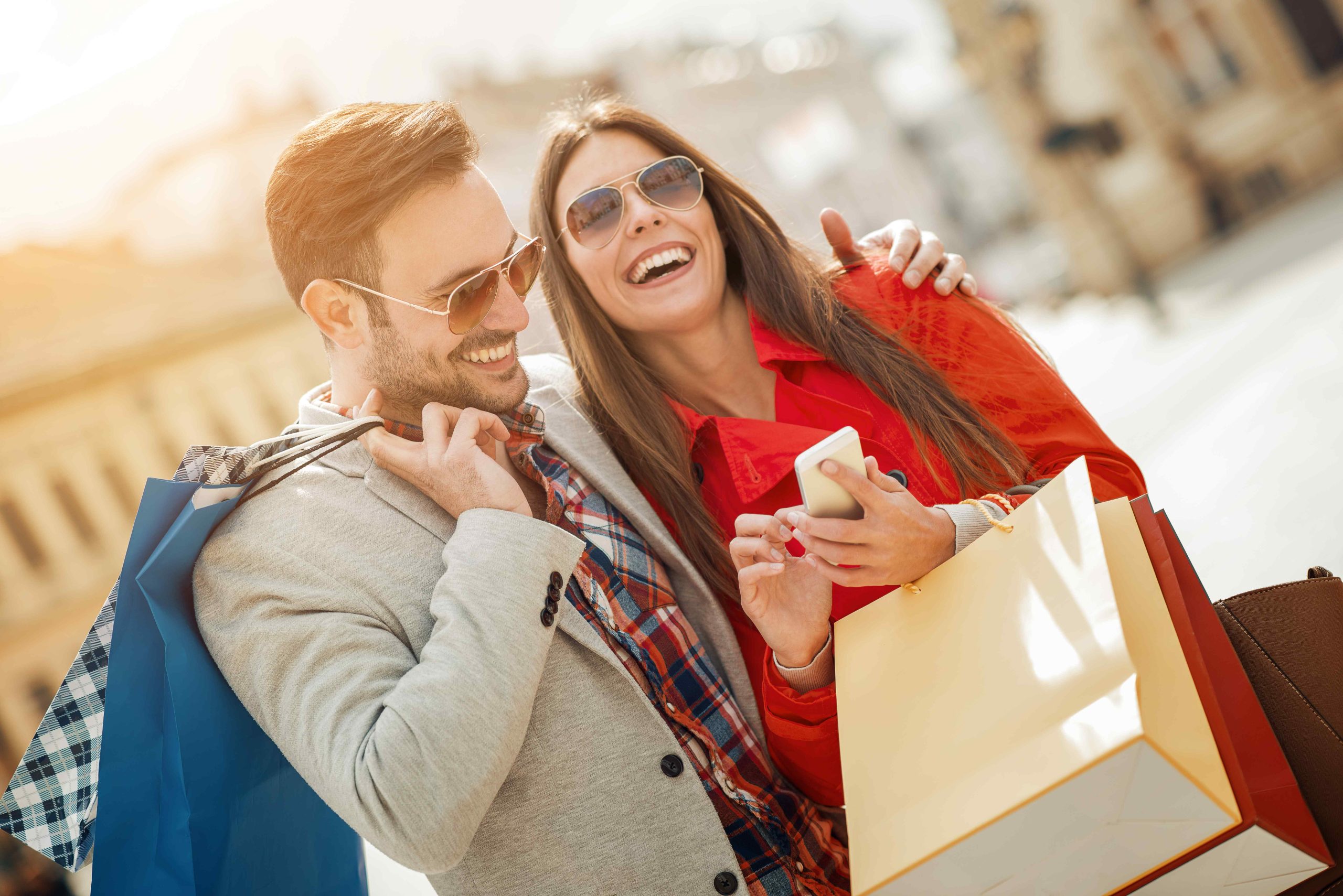 2 persons laughing and looking at a phone and next to this they are also holding two bags